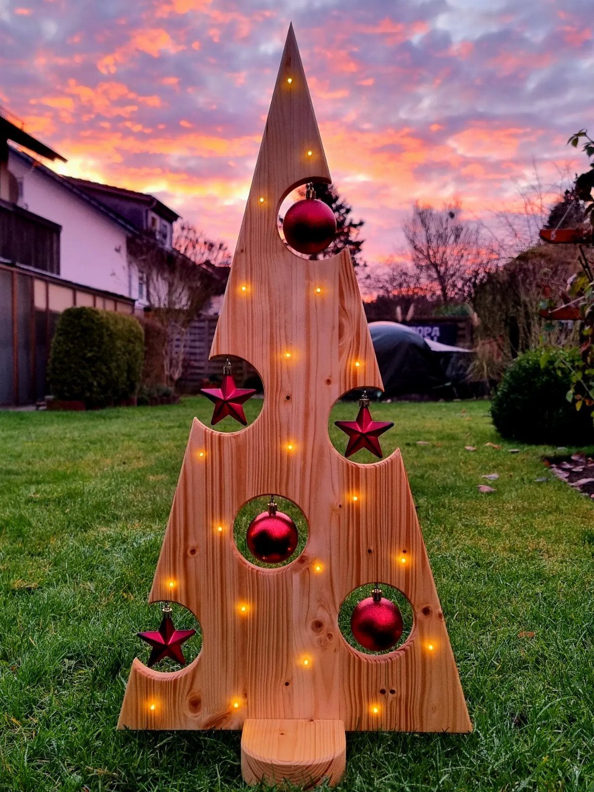 Wooden Christmas tree with red ornaments and warm lights on a green lawn