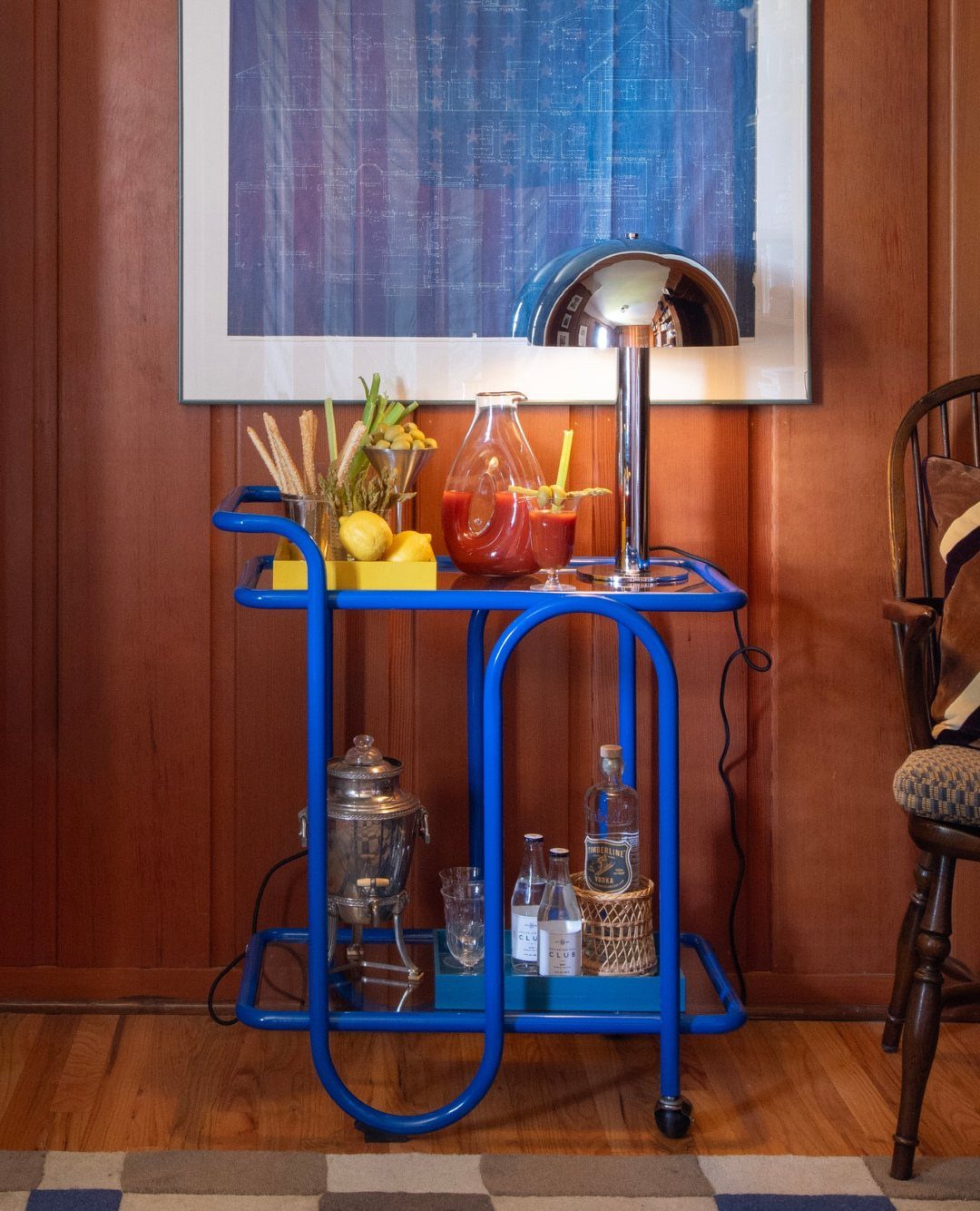 Blue tubular metal bar cart with drinks and a chrome lamp against wood paneling.
