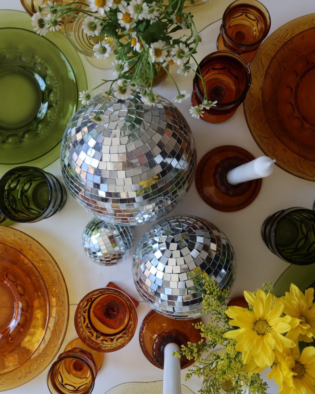 Two mirrored disco balls surrounded by amber and green glassware and yellow flowers.