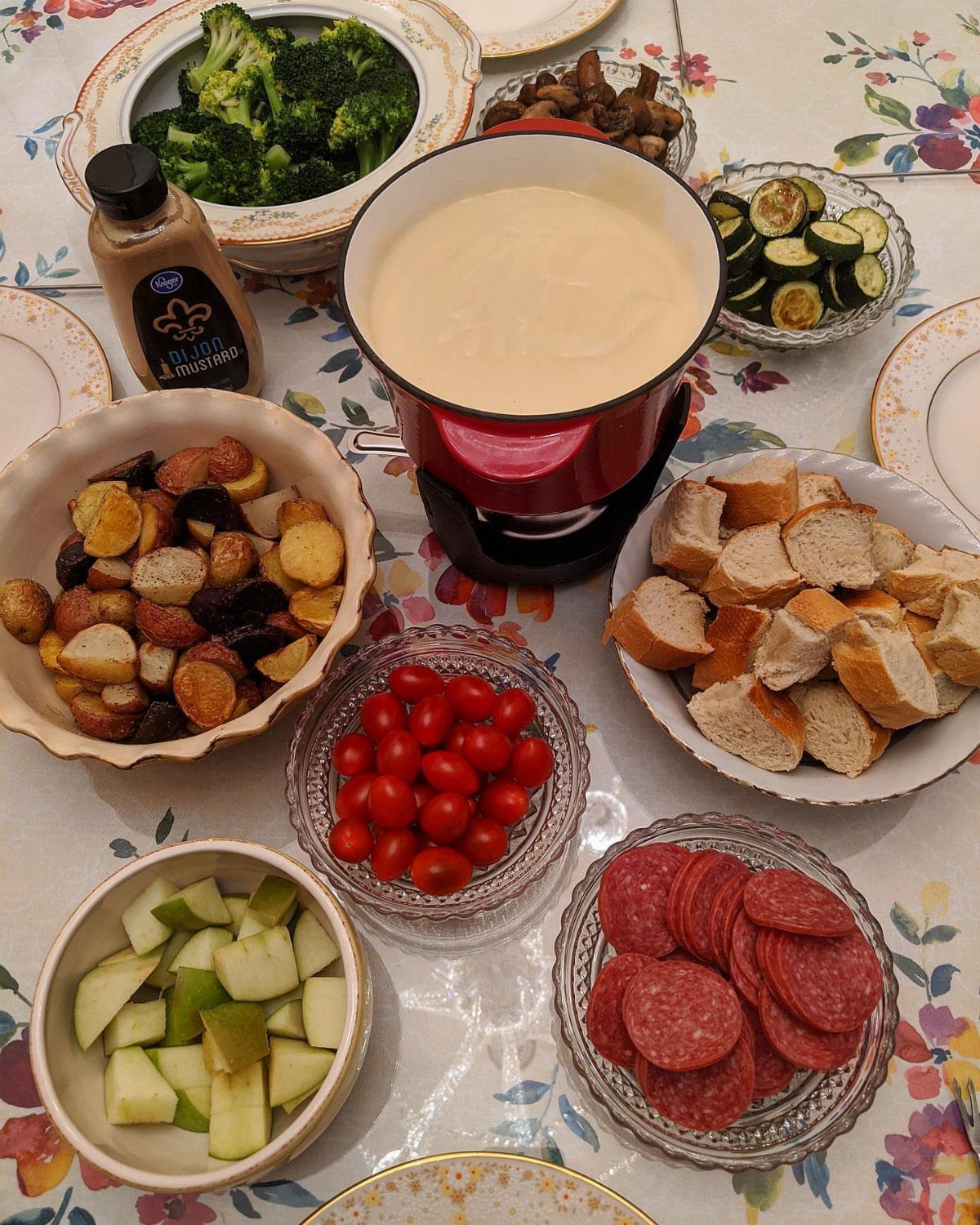 Red fondue pot with melted cheese surrounded by various dipping foods on floral tablecloth