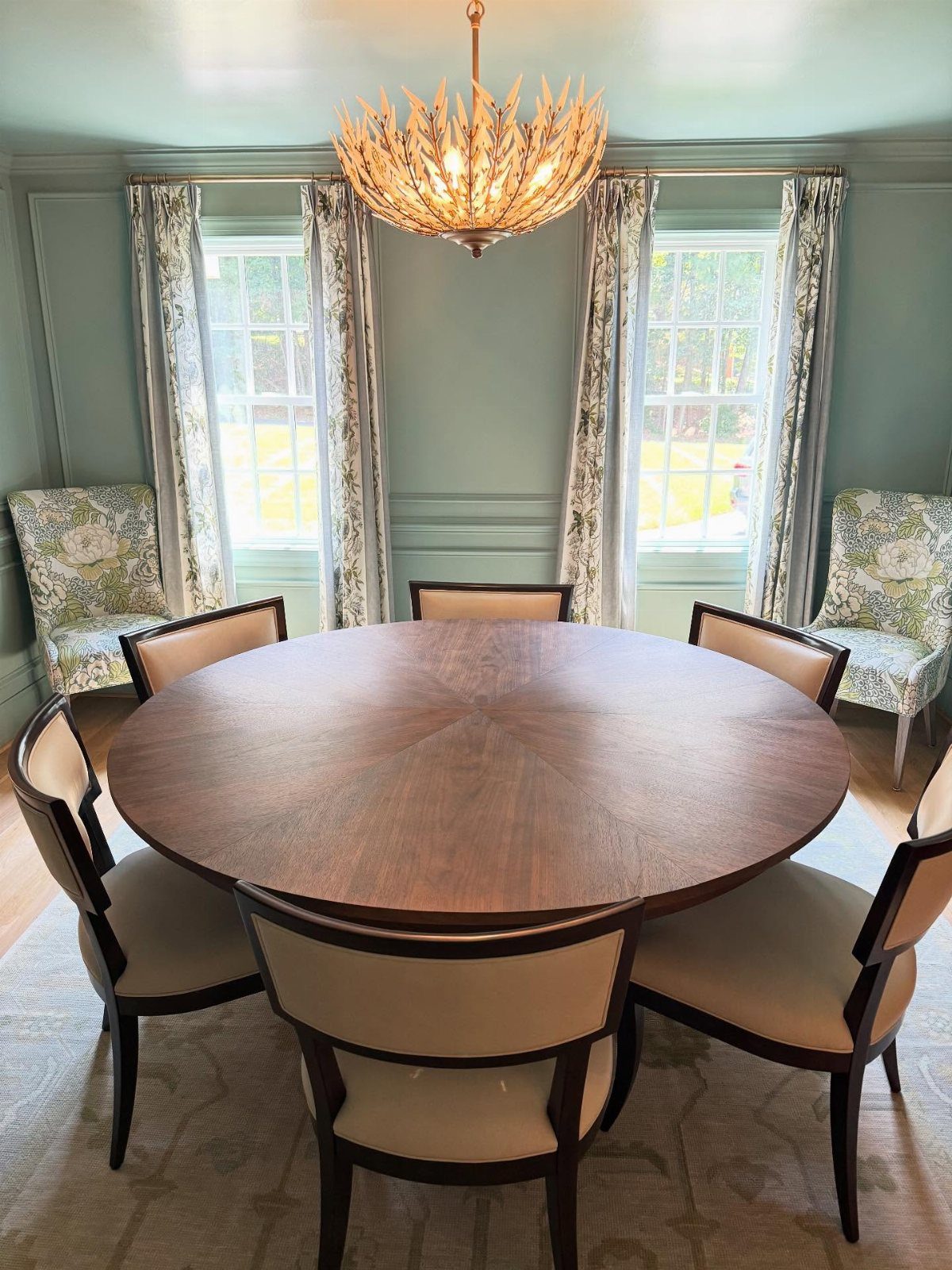 Round brown wooden dining table with six upholstered chairs under a gold chandelier