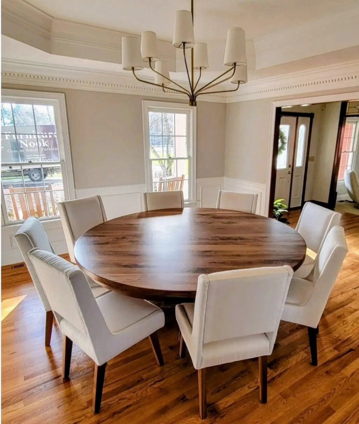 Round dark wood dining table with eight white upholstered chairs in a bright room.