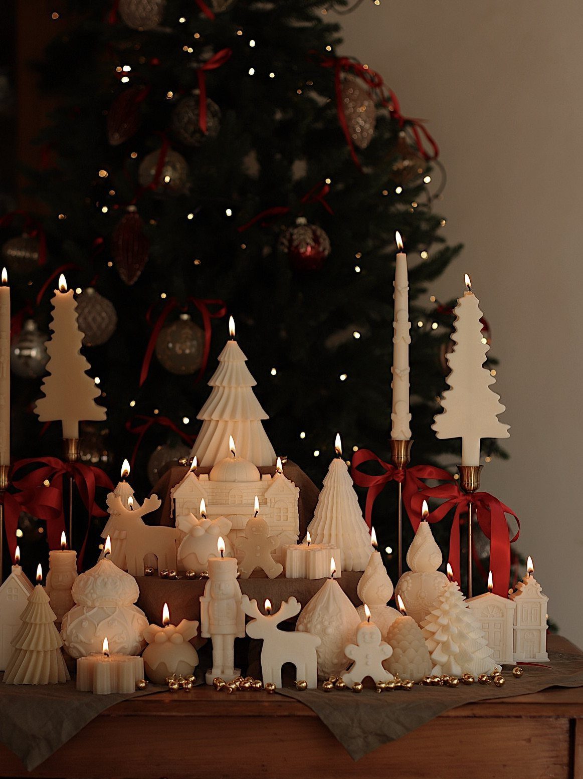 White Christmas-themed candles in various shapes lit on a wooden surface near a tree.