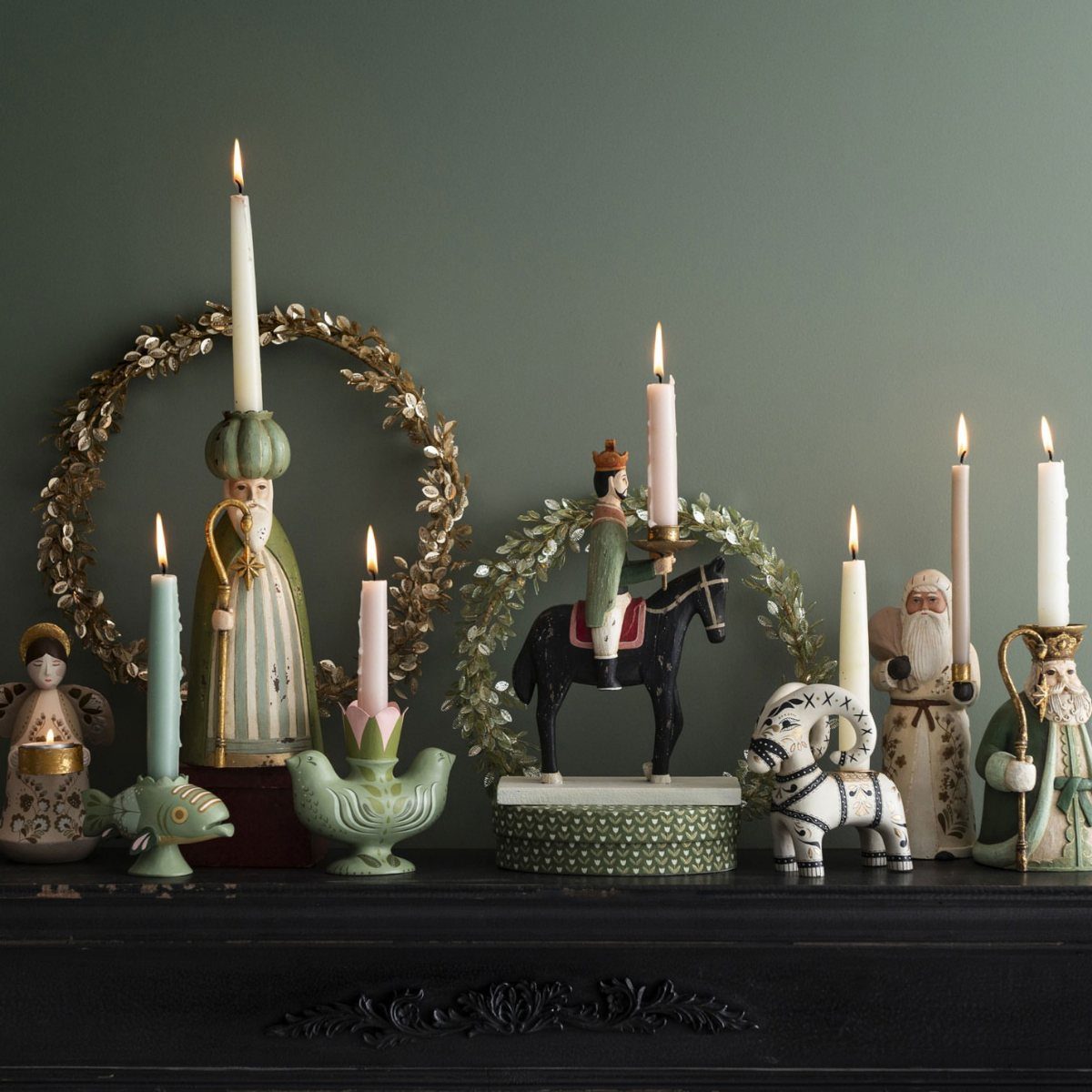 Lit taper candles in decorative figurines and wreaths on a black mantelpiece.