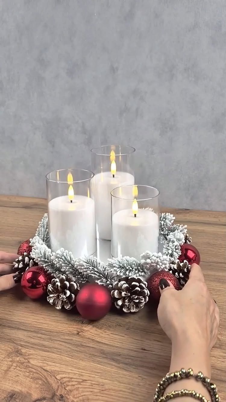 Three white candles in glass holders centered on a snowy holiday wreath with ornaments.