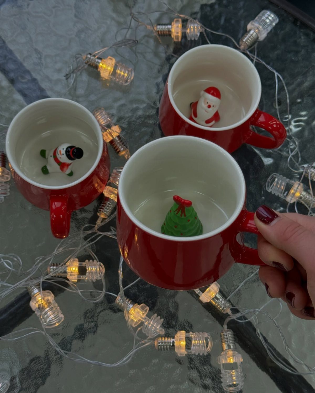 Three red mugs containing small Santa, snowman, and tree figurines among fairy lights.