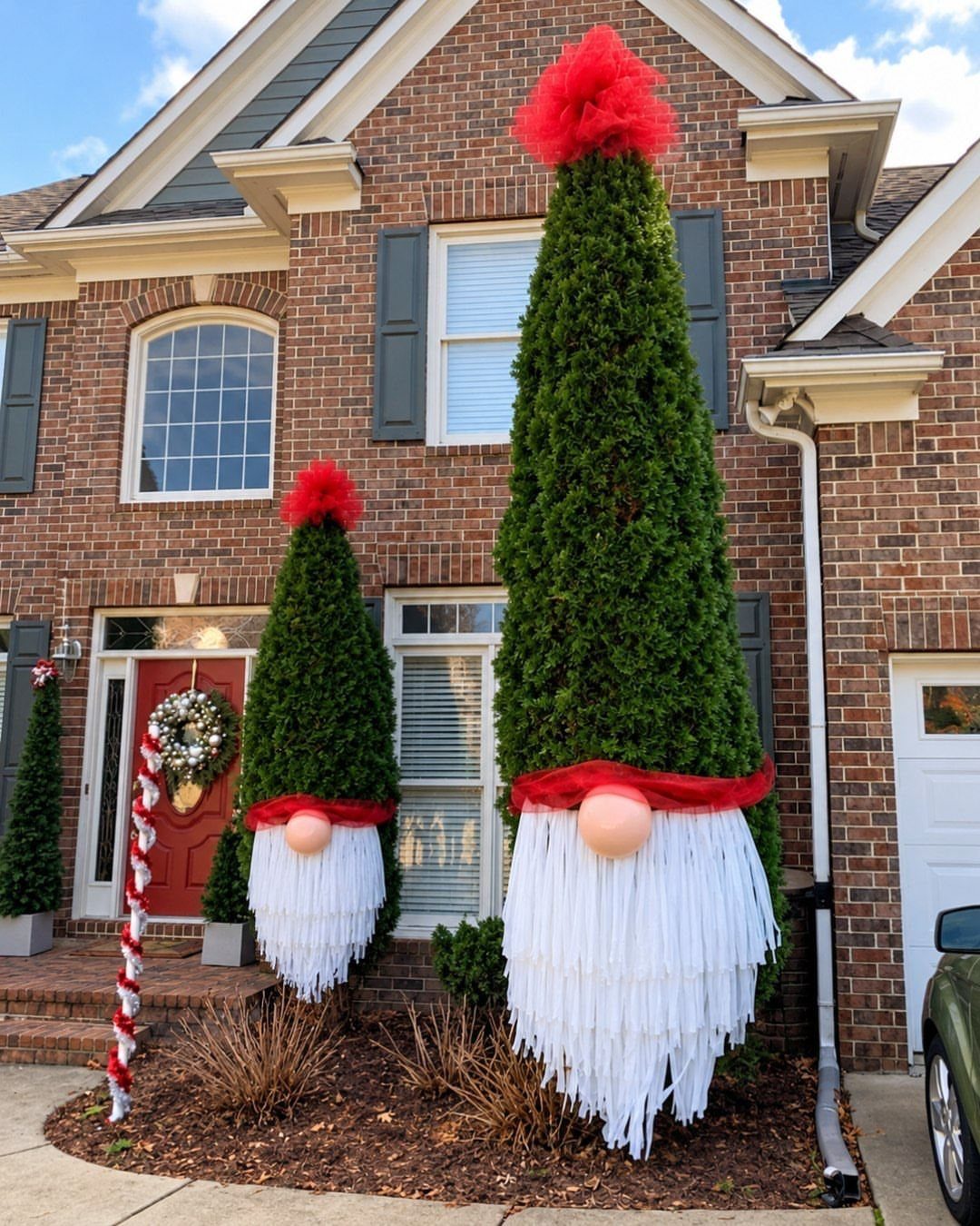 Two large topiary Christmas gnomes with red hats and white beards outside a house.