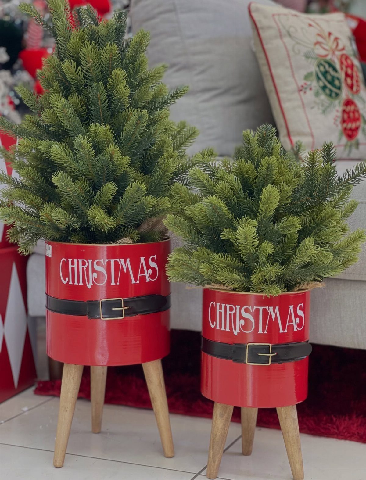 Two red Santa-themed planters with wooden legs holding small evergreen Christmas trees.