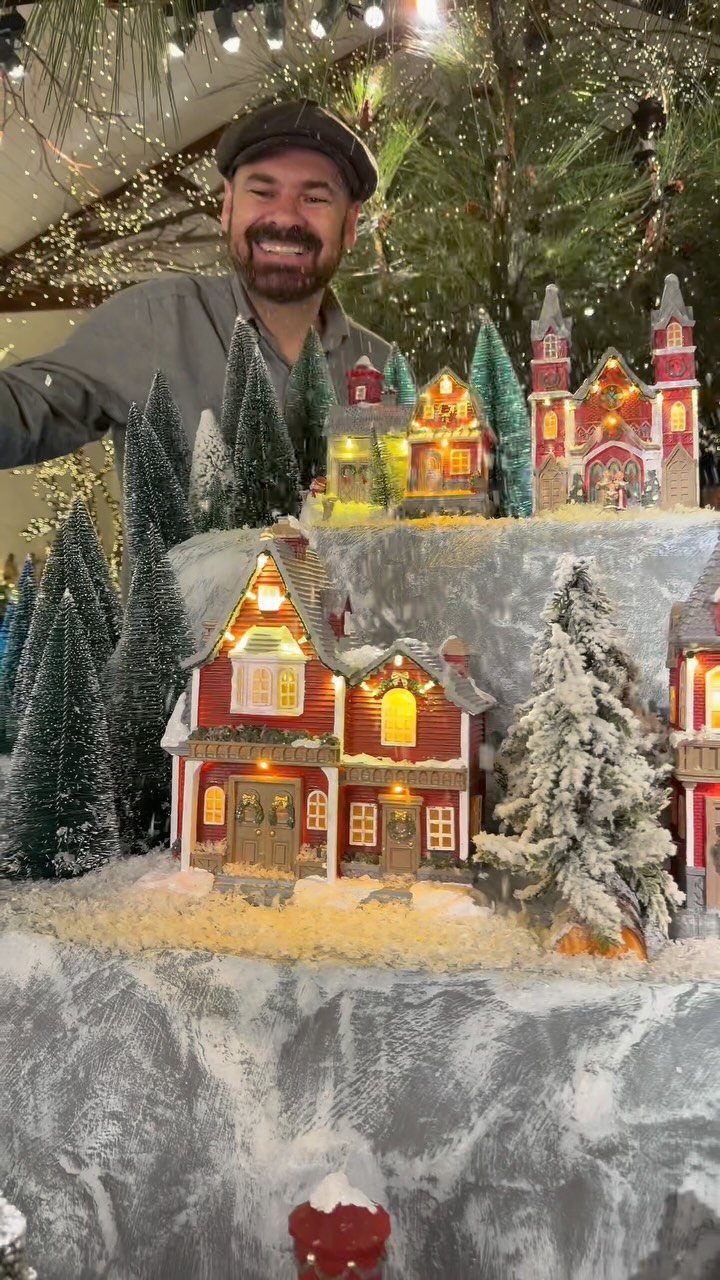Miniature winter village display with illuminated houses and snowy trees and a man.