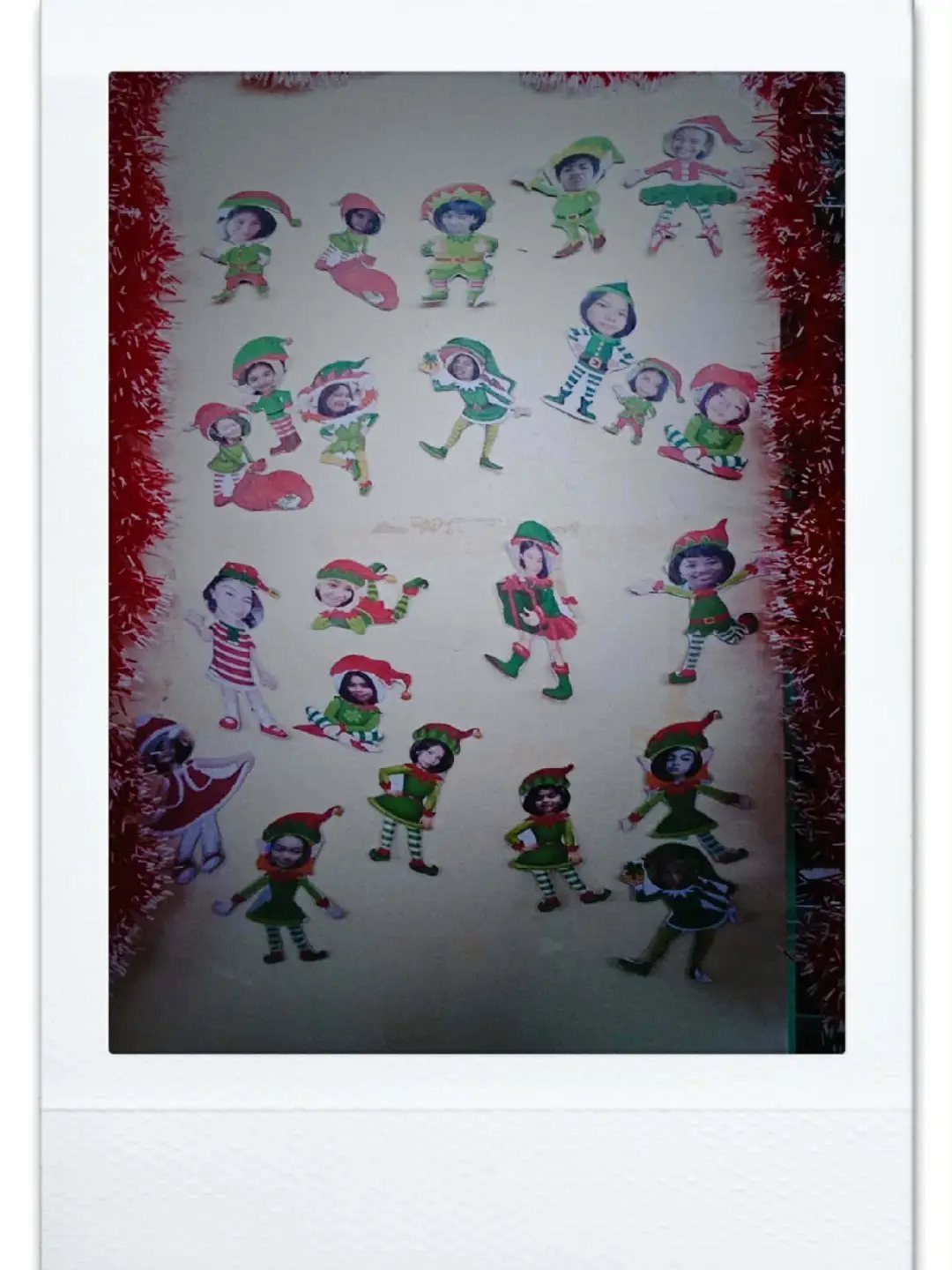 Elf cutouts with human faces pasted on them arranged on a white wall.