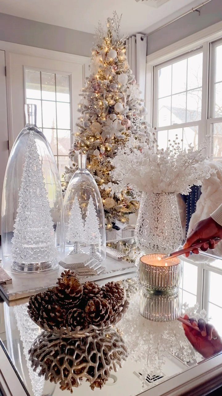 Mirrored coffee table with Christmas cloches, pinecones, and a candle near a flocked tree.
