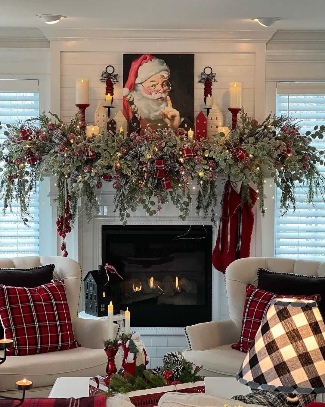Decorated fireplace mantel with Santa portrait, festive greenery, and red plaid pillows.