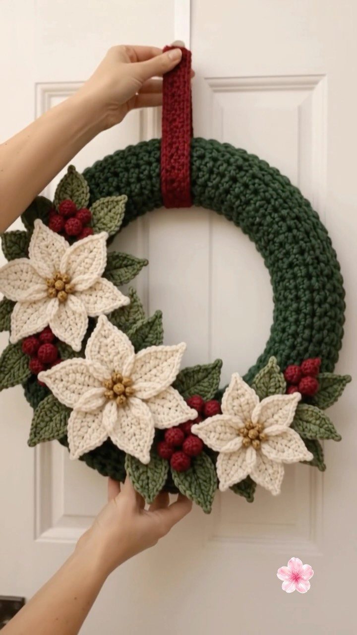 Crocheted green wreath with cream poinsettias and red berries hanging on a white door