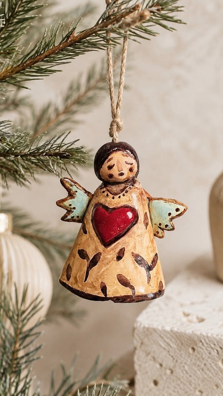 Handmade ceramic angel ornament with a red heart hanging from a pine branch