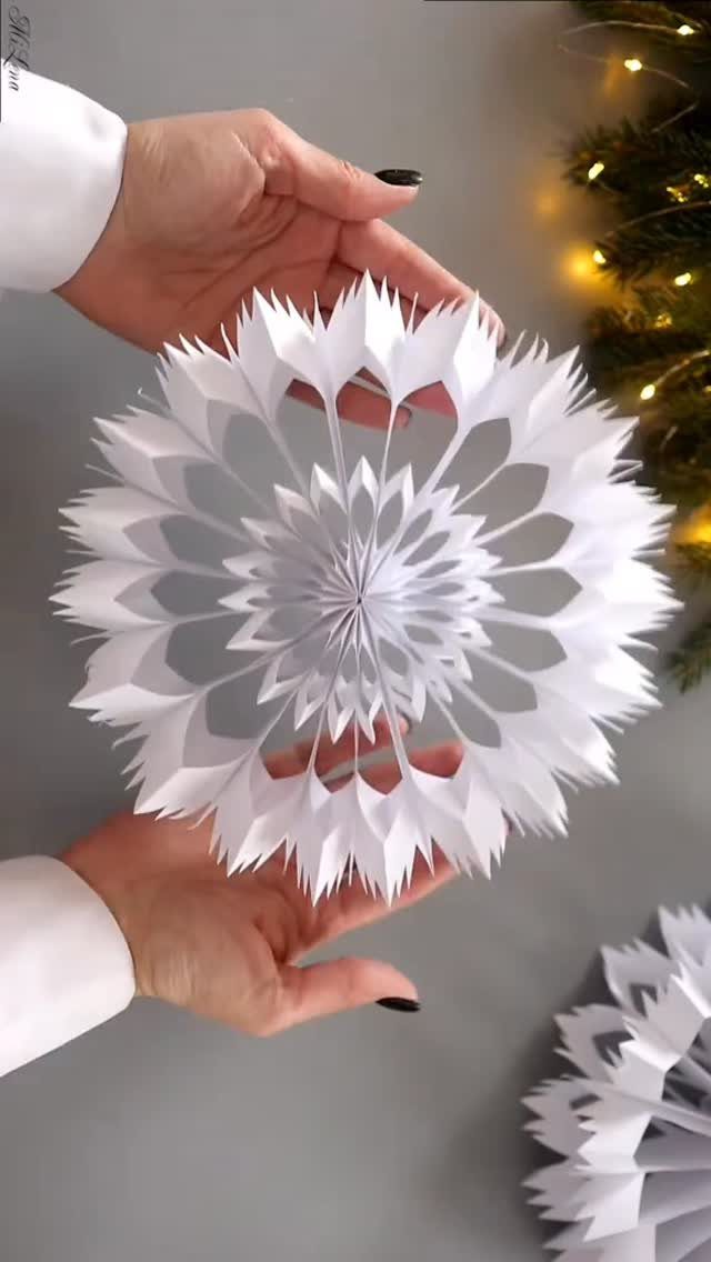 White 3D paper snowflake held by hands against a grey background with lights.