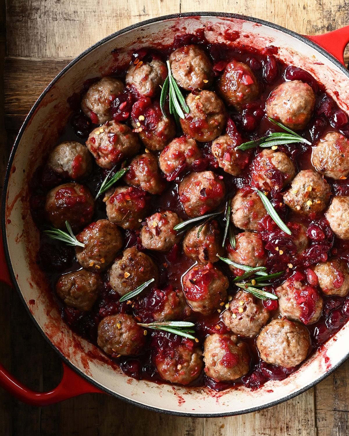 Meatballs in red cranberry sauce with rosemary sprigs in a red cast iron pan.