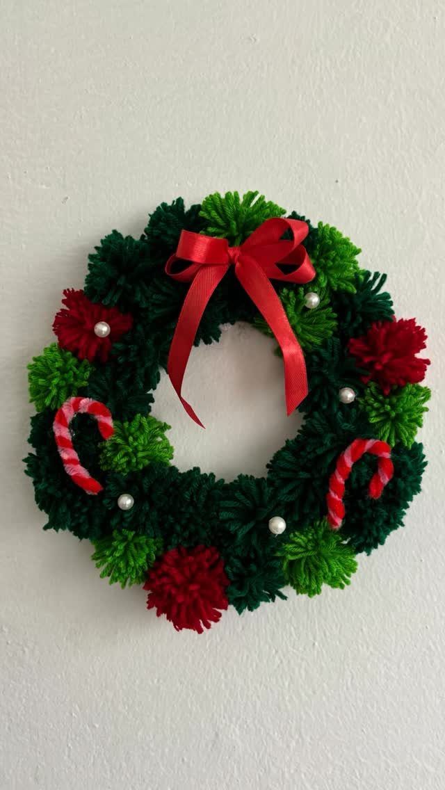 Yarn pom-pom Christmas wreath with red bow and candy canes on white wall