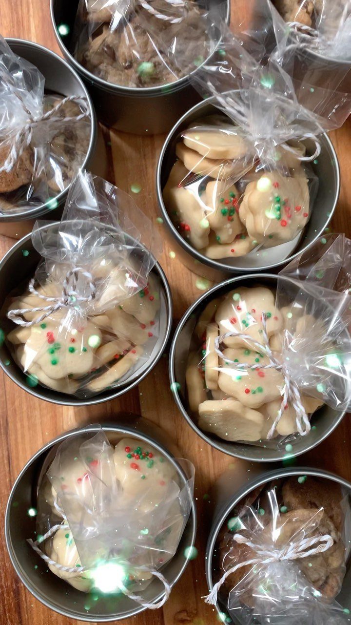 Cookies with festive sprinkles in clear bags inside round metal tins on wood.