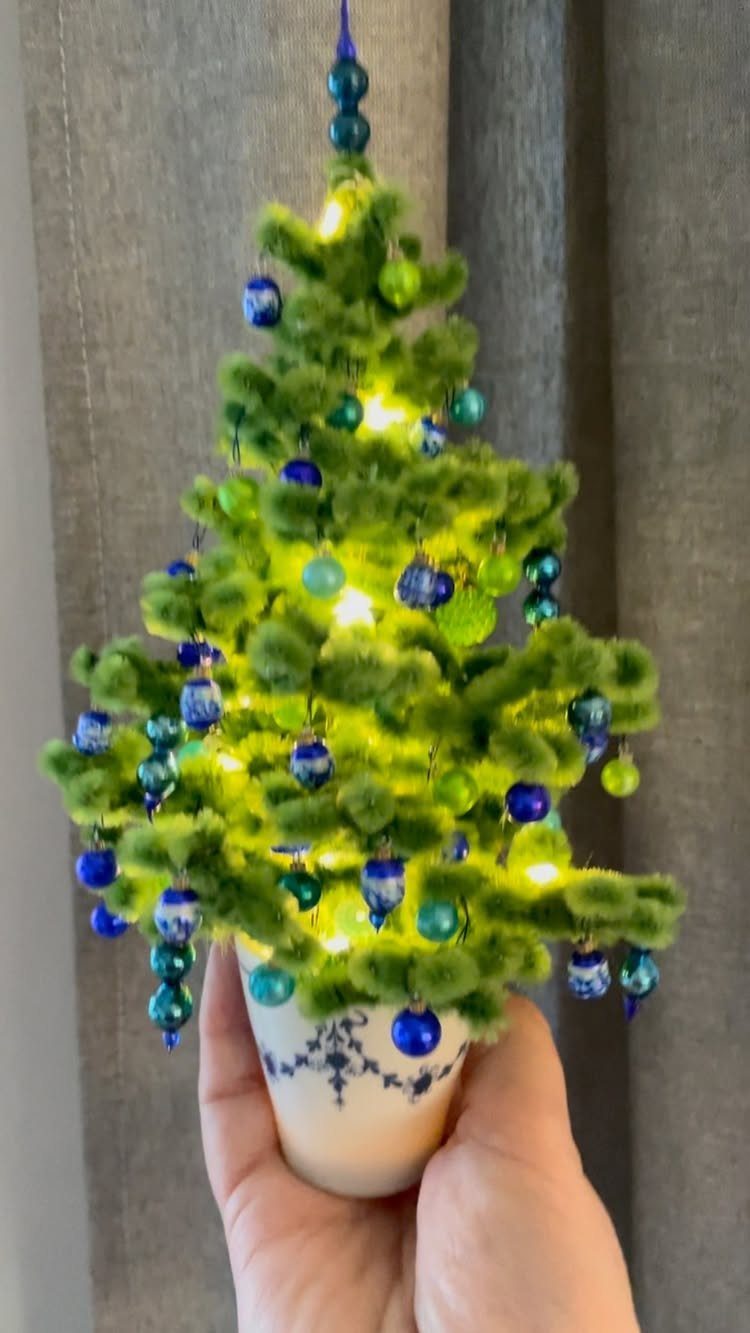 Small fluffy green Christmas tree with blue ornaments and lights in a white pot.