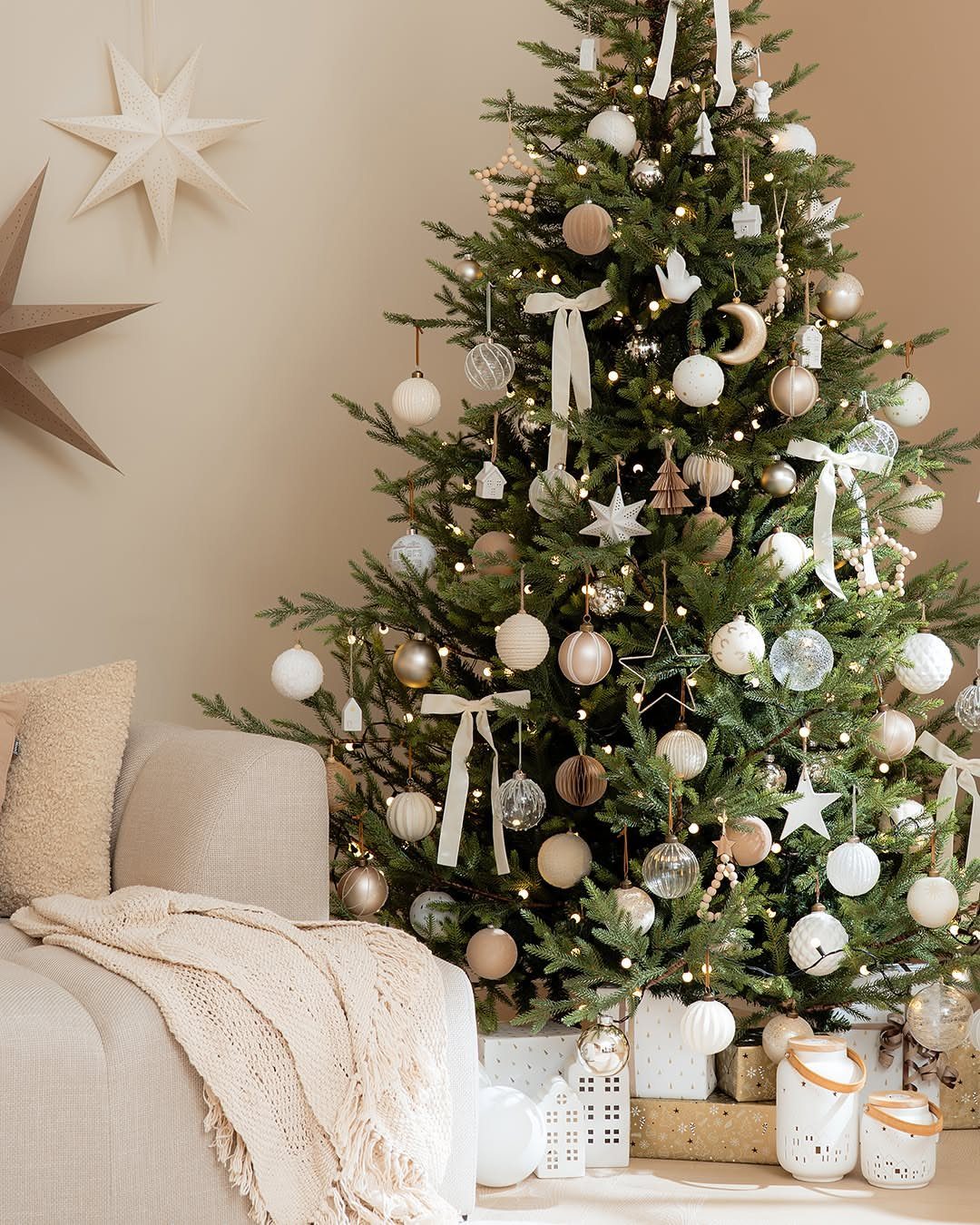 Decorated Christmas tree with neutral ornaments and white ribbons beside a cozy beige sofa.