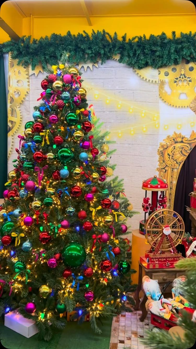 Colorful Christmas tree decorated with rainbow ornaments and lights against a white brick wall.