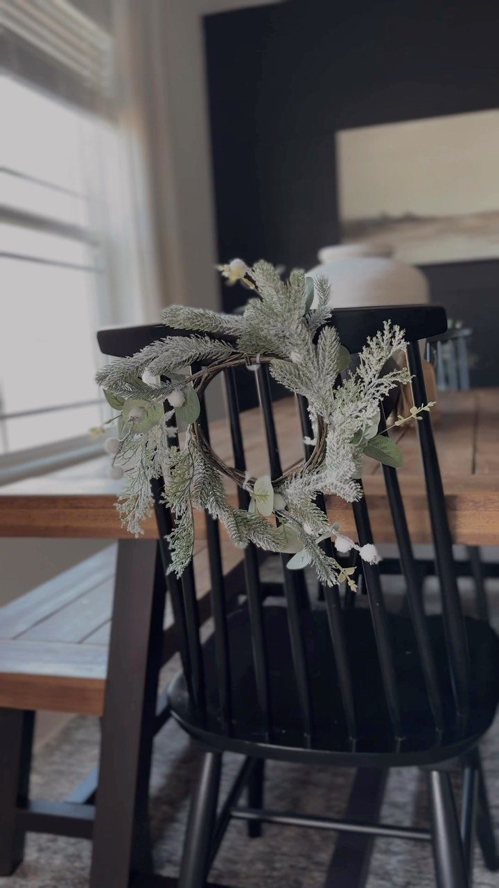 Frosted evergreen wreath hanging on the back of a black wooden dining chair.