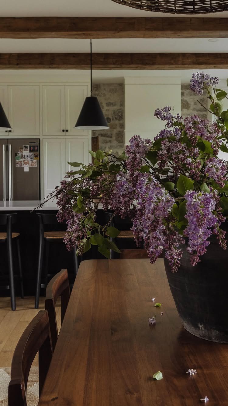Wooden dining table with purple lilacs in a vase and dark wooden chairs.