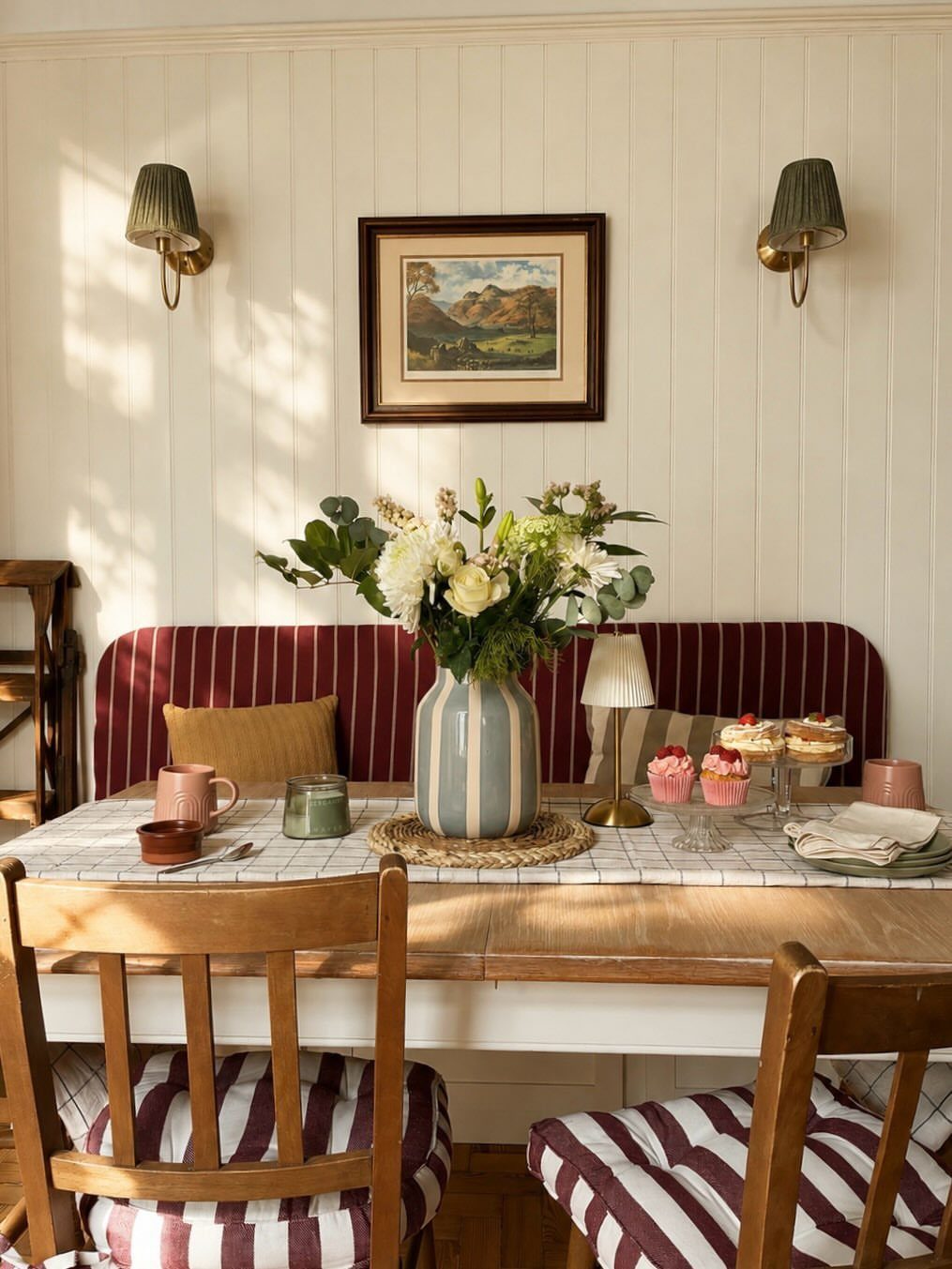 Wooden dining table with striped seating and white paneled walls in a farmhouse style.