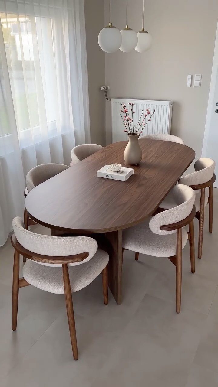 Oval wooden dining table with beige chairs placed in a bright room corner.