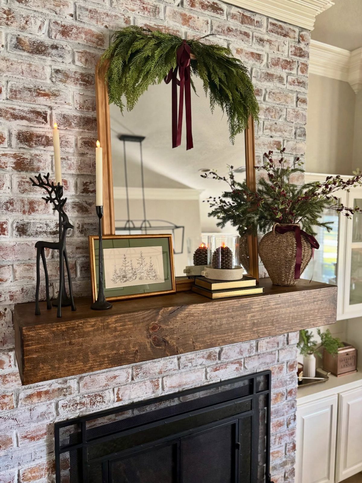 Wooden mantel decorated asymmetrically with greenery, candles, and a mirror on brick wall.
