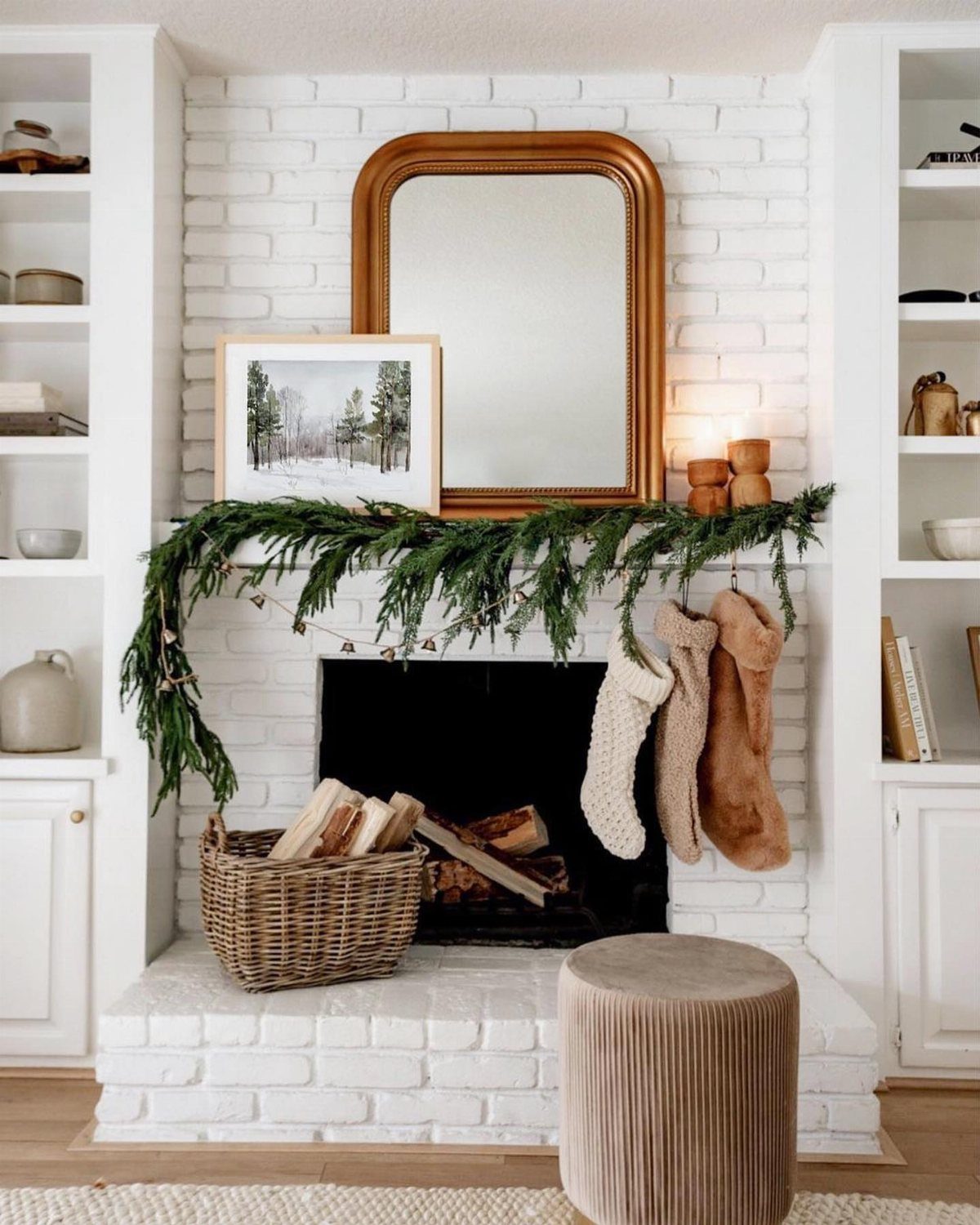 White brick fireplace mantel decorated with green garland, neutral stockings, and gold mirror.