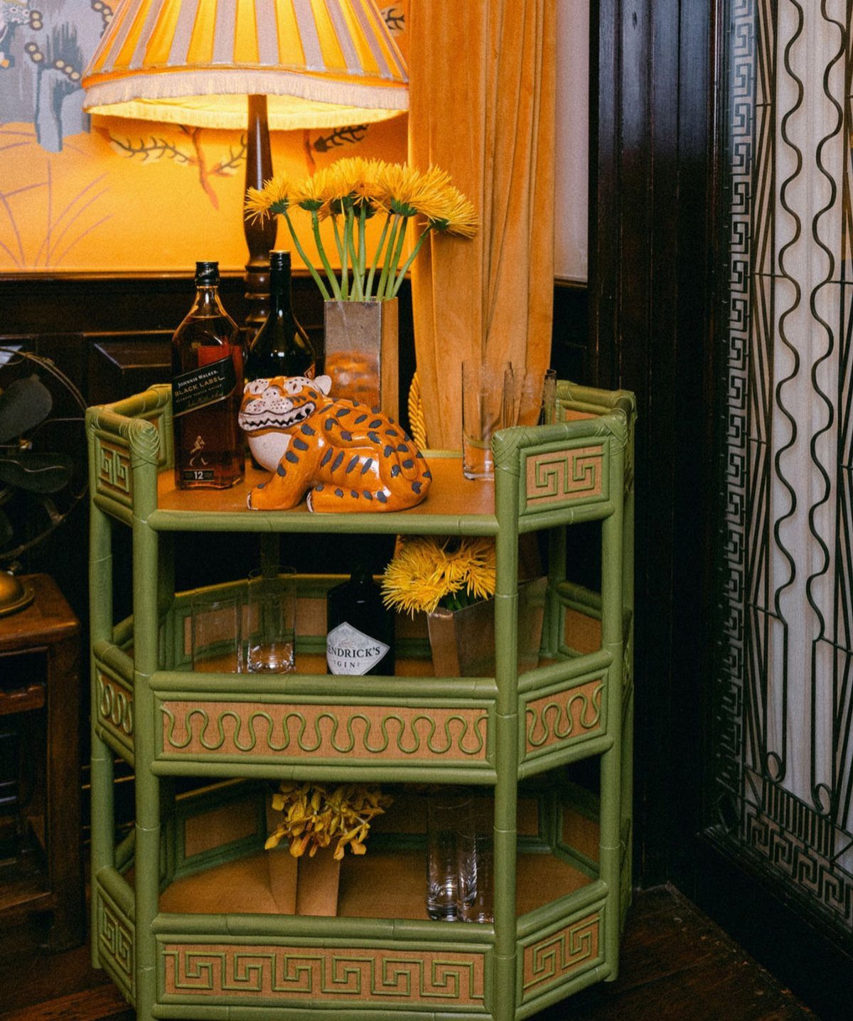 Green bamboo bar cart with liquor bottles, yellow flowers, and a tiger figurine.