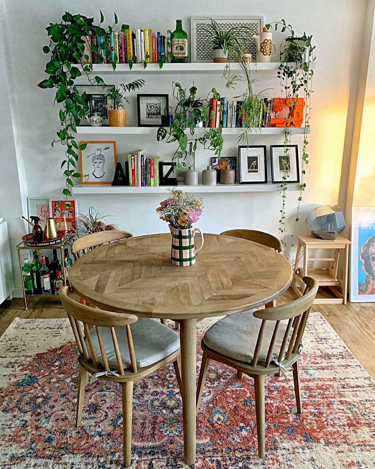 Round wooden dining table with chairs, featuring wall shelves with plants and artwork.