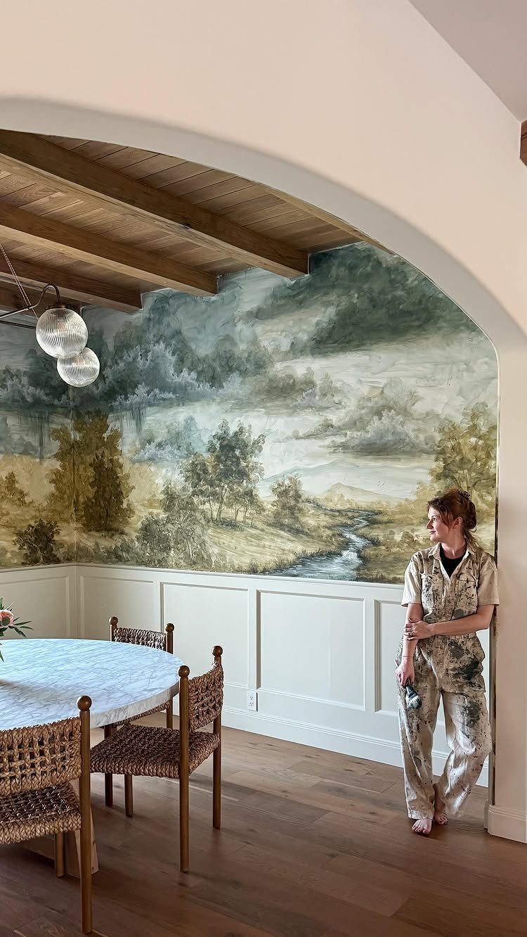 Landscape wall mural in a dining room with white wainscoting and woven chairs.