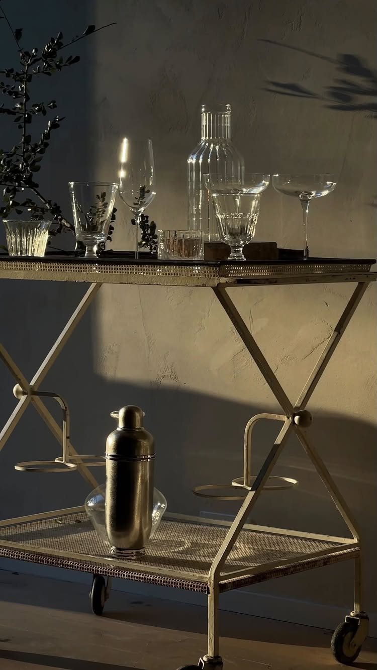 Gold metal bar cart with glassware and a cocktail shaker against a neutral wall.