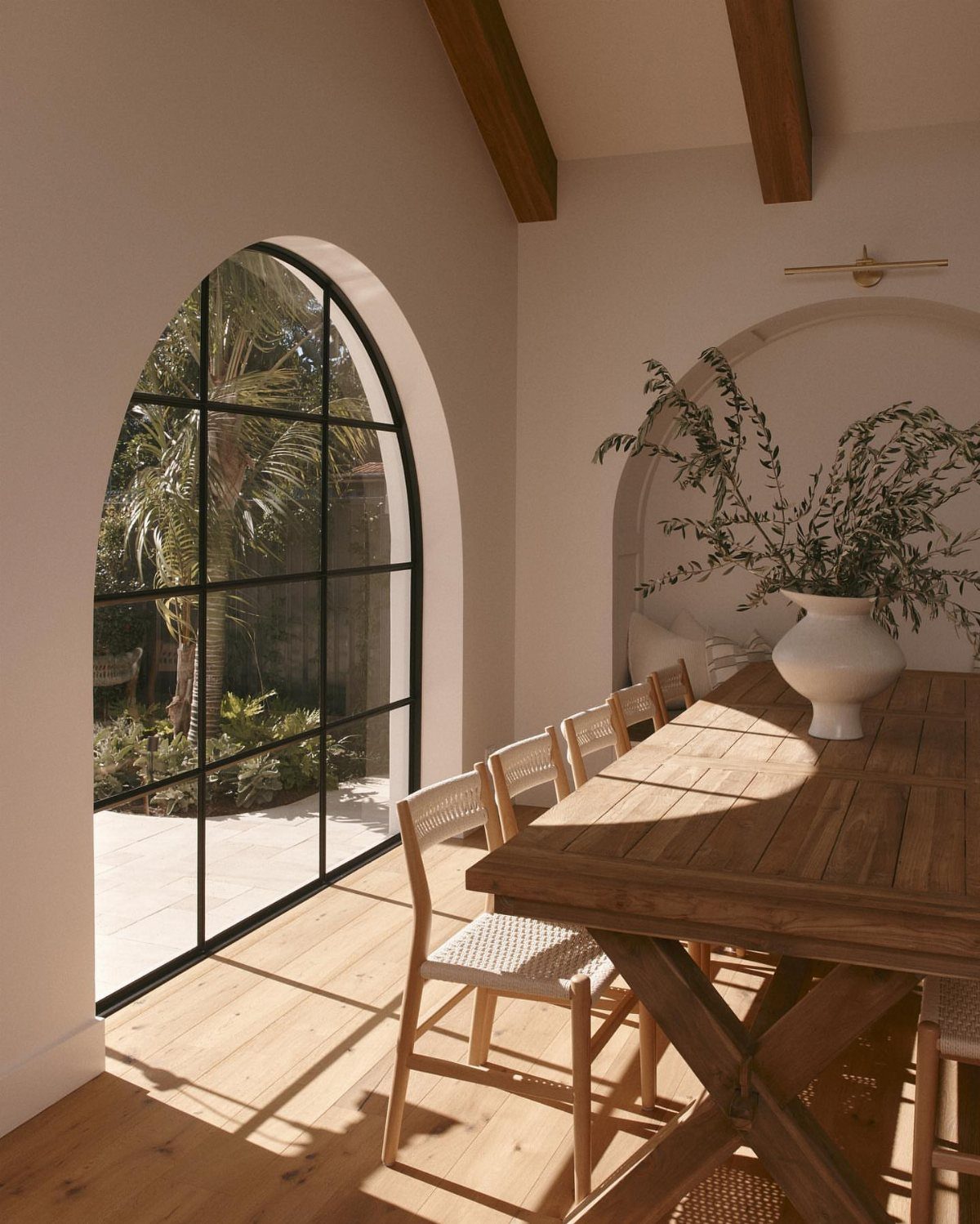 Wooden dining table and chairs placed beside a large arched window with greenery.