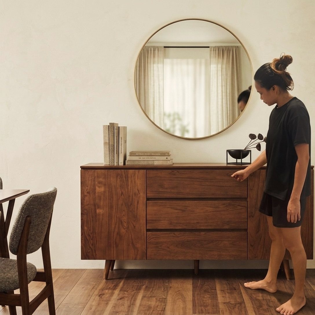 Wooden buffet with a round gold mirror and books against a white wall.