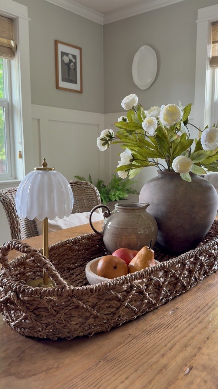 Woven wicker tray on wooden table with fruit, pitcher, and flower vase.