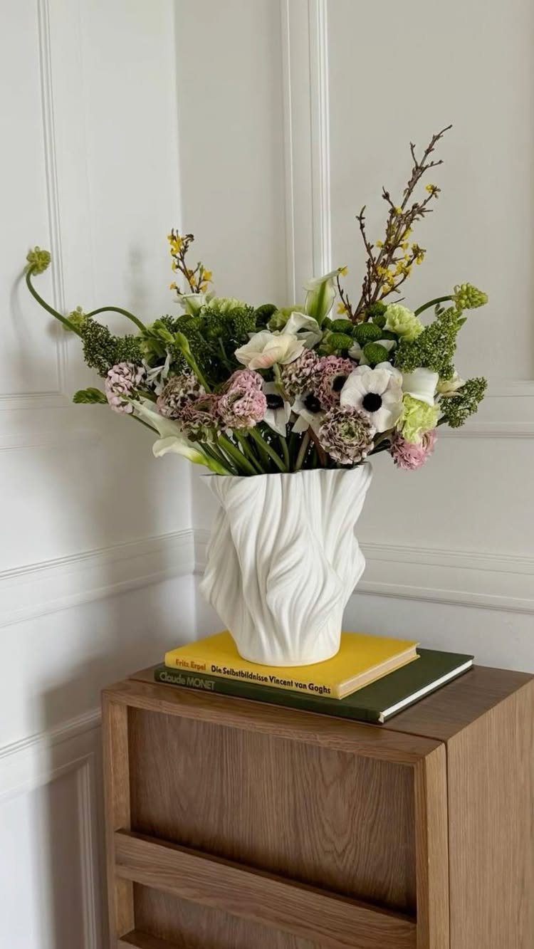 White sculptural vase with flowers resting on books atop a wooden cabinet.