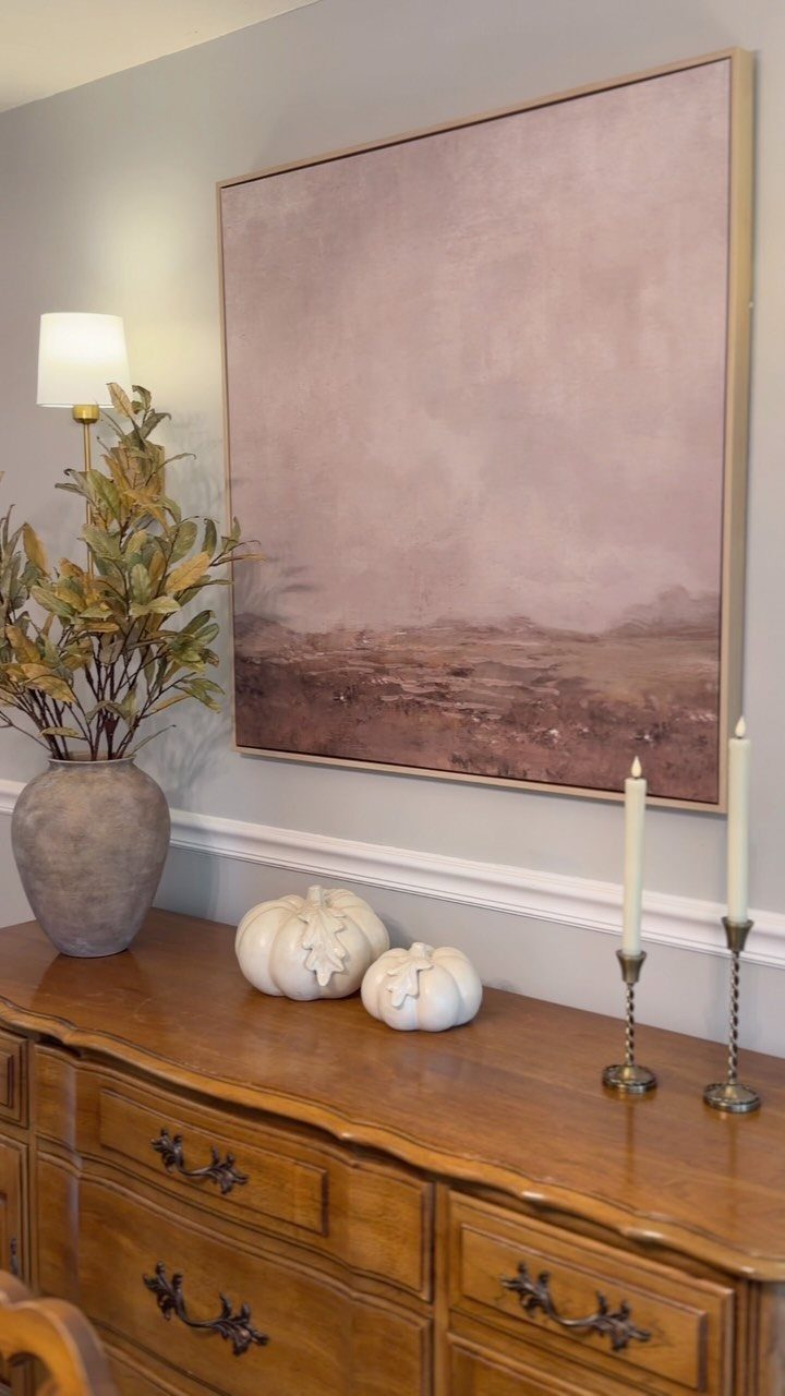 Wooden buffet decorated with white pumpkins, candles, and foliage under an abstract painting.