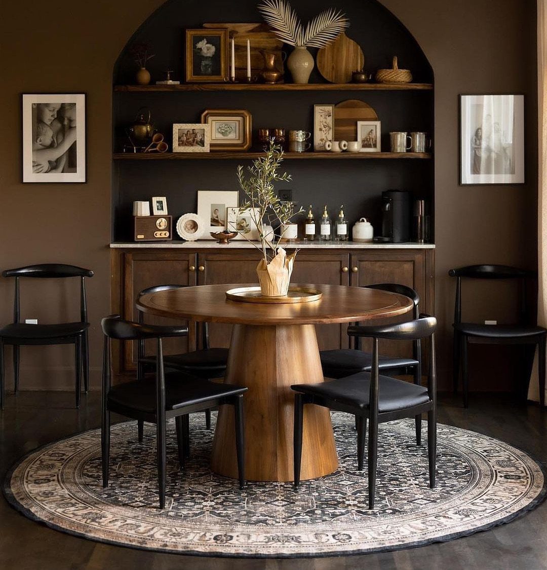Round wooden dining table with black chairs in a dark brown room with alcove