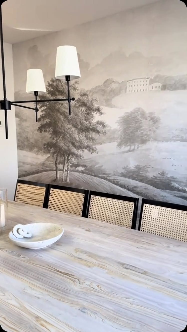 Light wood dining table with cane chairs against a monochrome landscape wall mural.