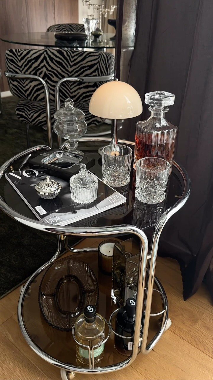 Chrome bar cart with black glass shelves holding a decanter and mushroom lamp.