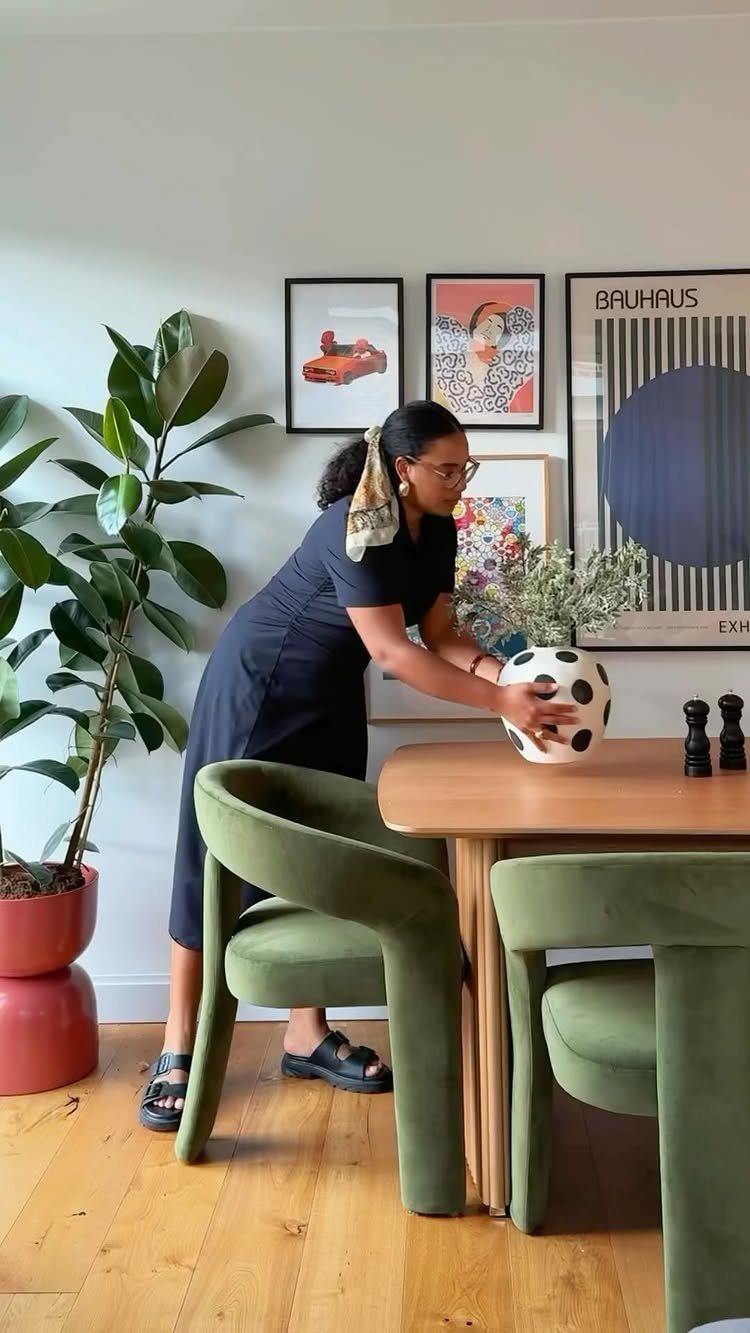 Woman arranging a vase on a wooden table with green velvet chairs and plants.