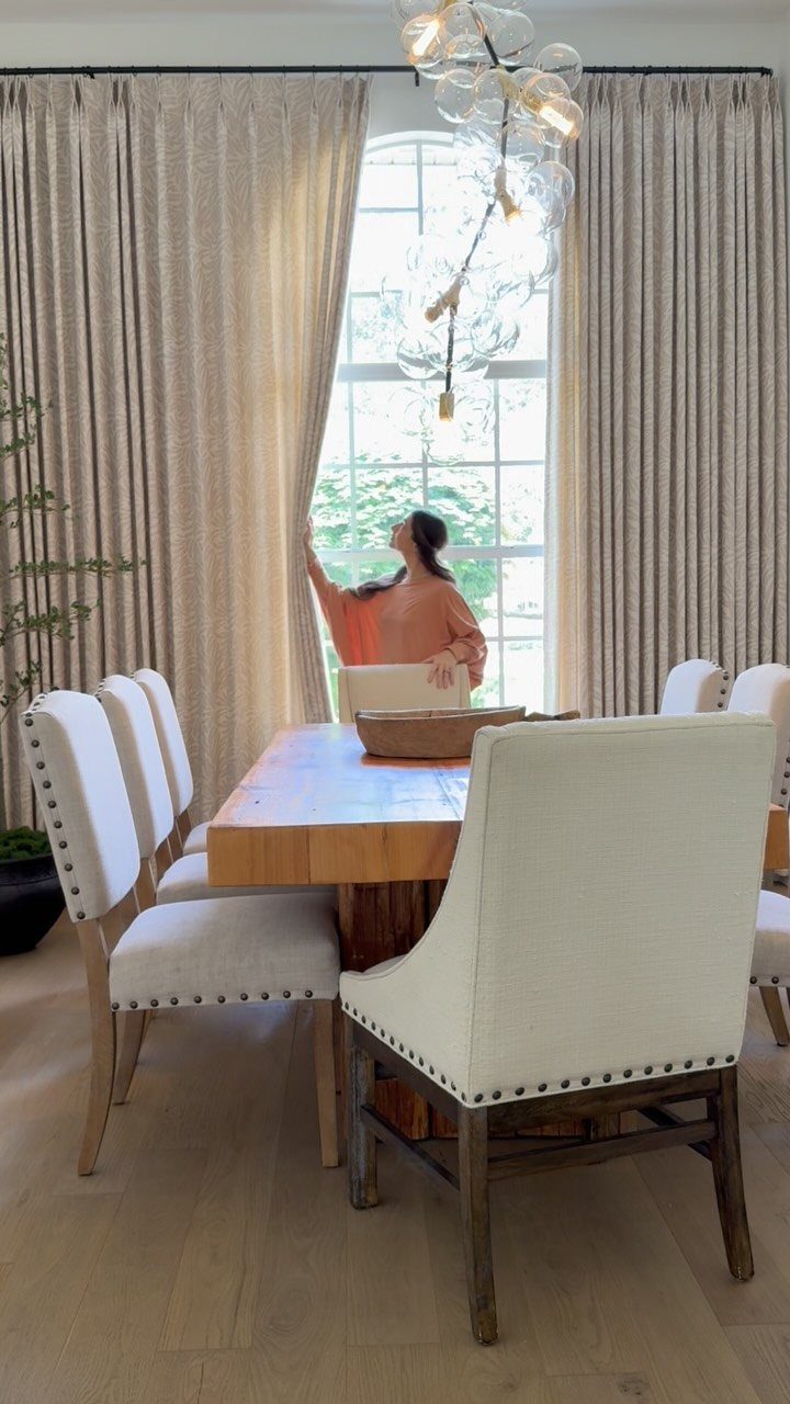 Beige curtains framing a window in a dining room with white upholstered chairs.