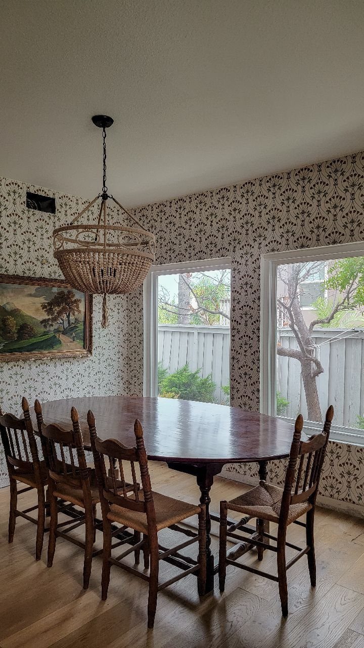Dining room with black and white floral wallpaper, oval wooden table and wicker chandelier