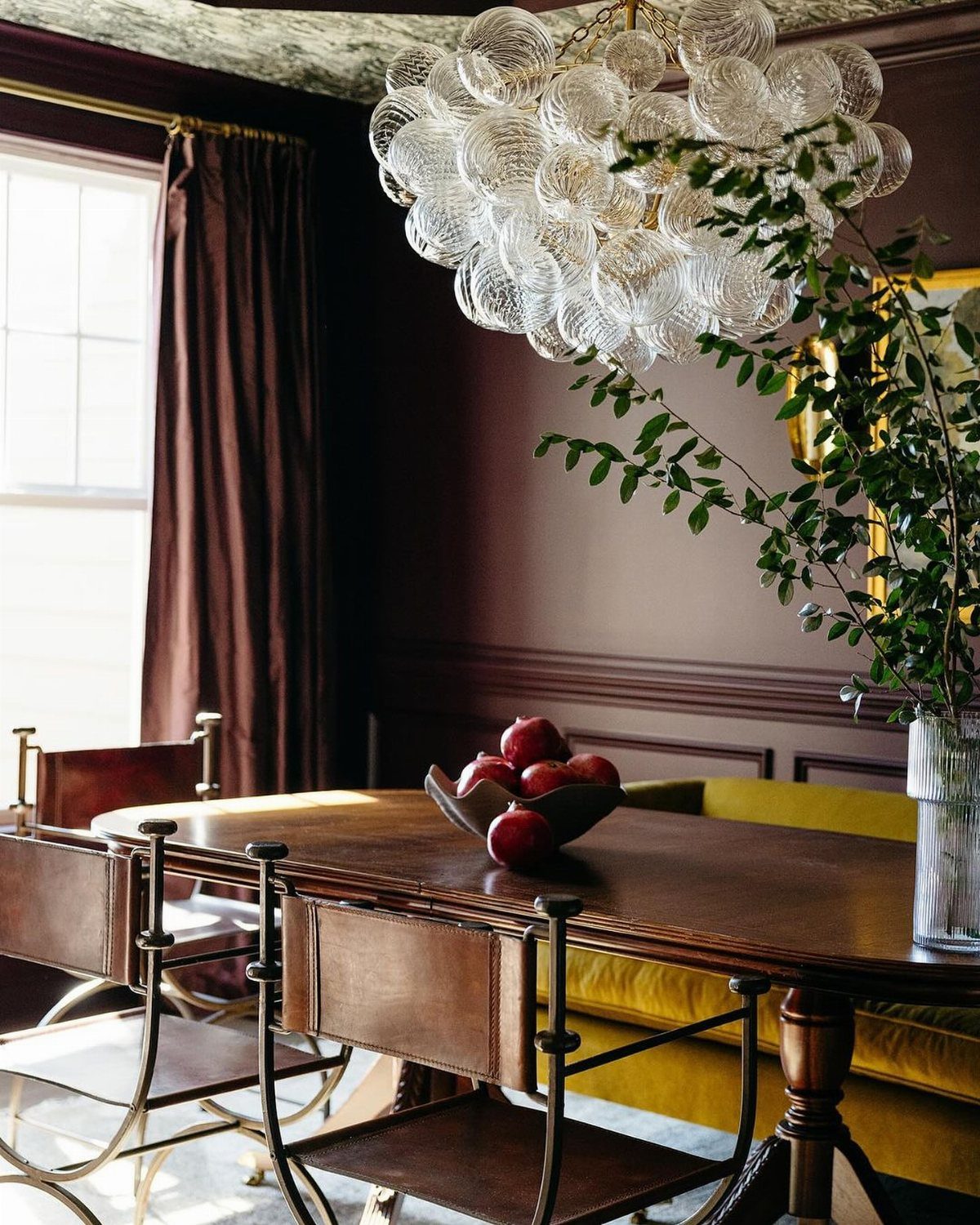 Wooden dining table with red apples, yellow seating, and burgundy walls under a bubble chandelier.