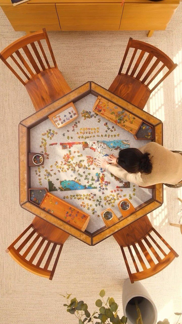 Hexagonal wooden dining table with four chairs and a person doing a puzzle.