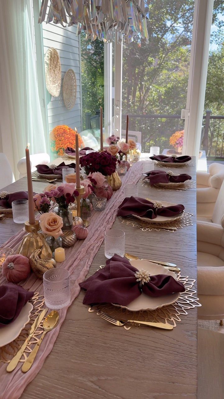 Wooden dining table with low floral centerpieces, burgundy napkins, and gold place settings.