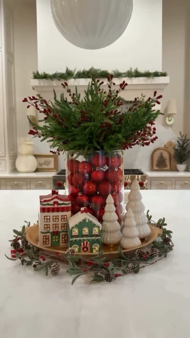 Wooden tray centerpiece with red ornaments in a vase, miniature houses, and white trees.